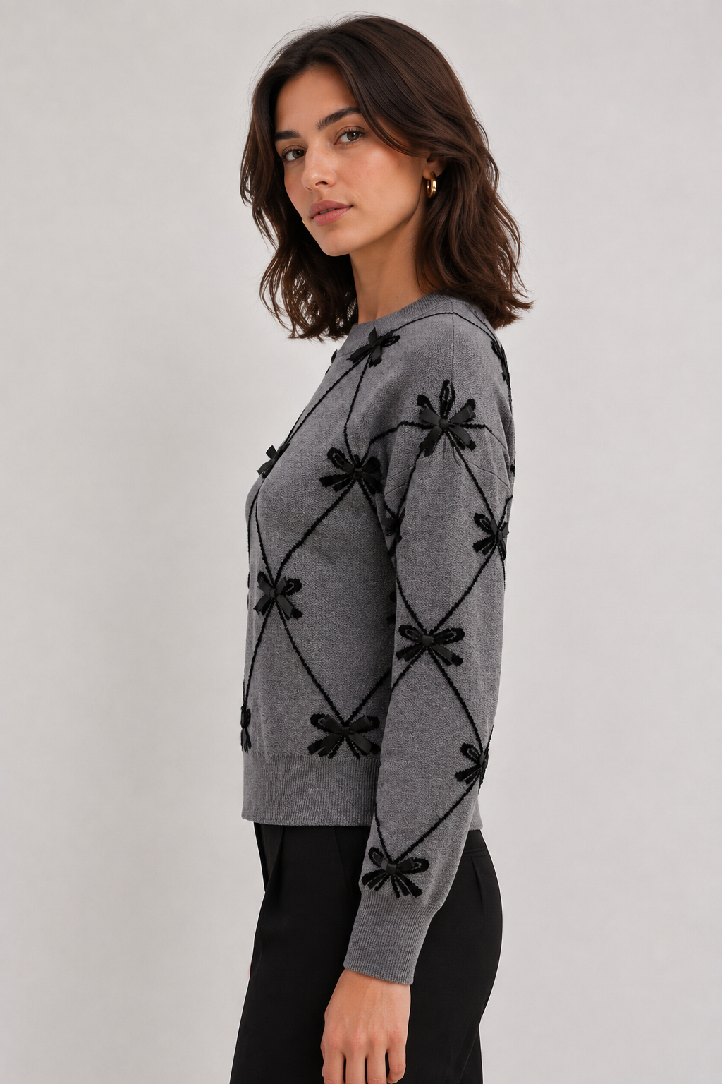 Point Zero grey sweater front view on a woman, showing the black diamond lattice pattern with bow embellishments and crew neckline"